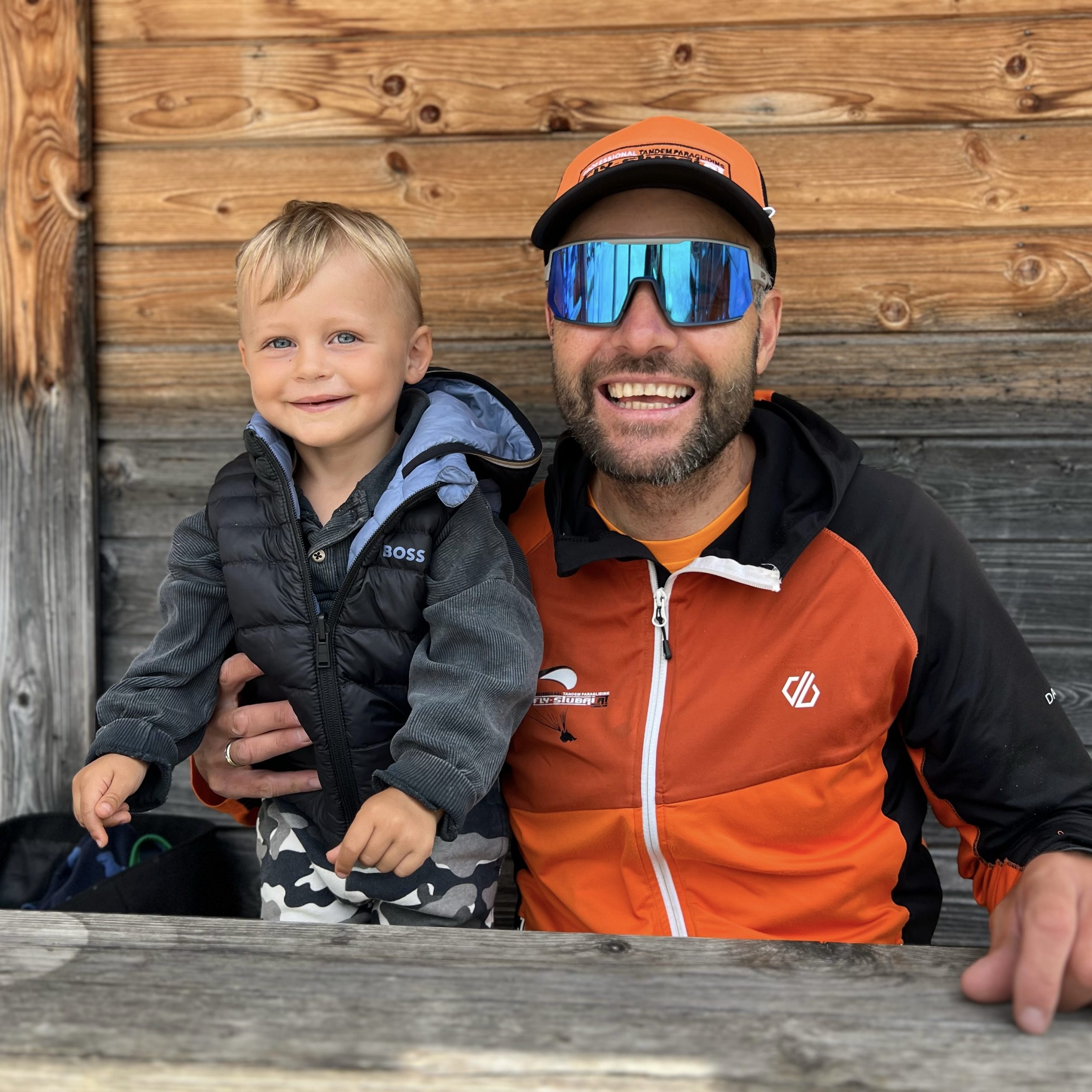 Ein lächelnder Mann in orangefarbener Sportkleidung und reflektierender Sonnenbrille sitzt neben einem kleinen Jungen in schwarzer Weste und grauer Tarnhose. Sie sitzen an einem rustikalen Holztisch mit einer Holzwand im Hintergrund.