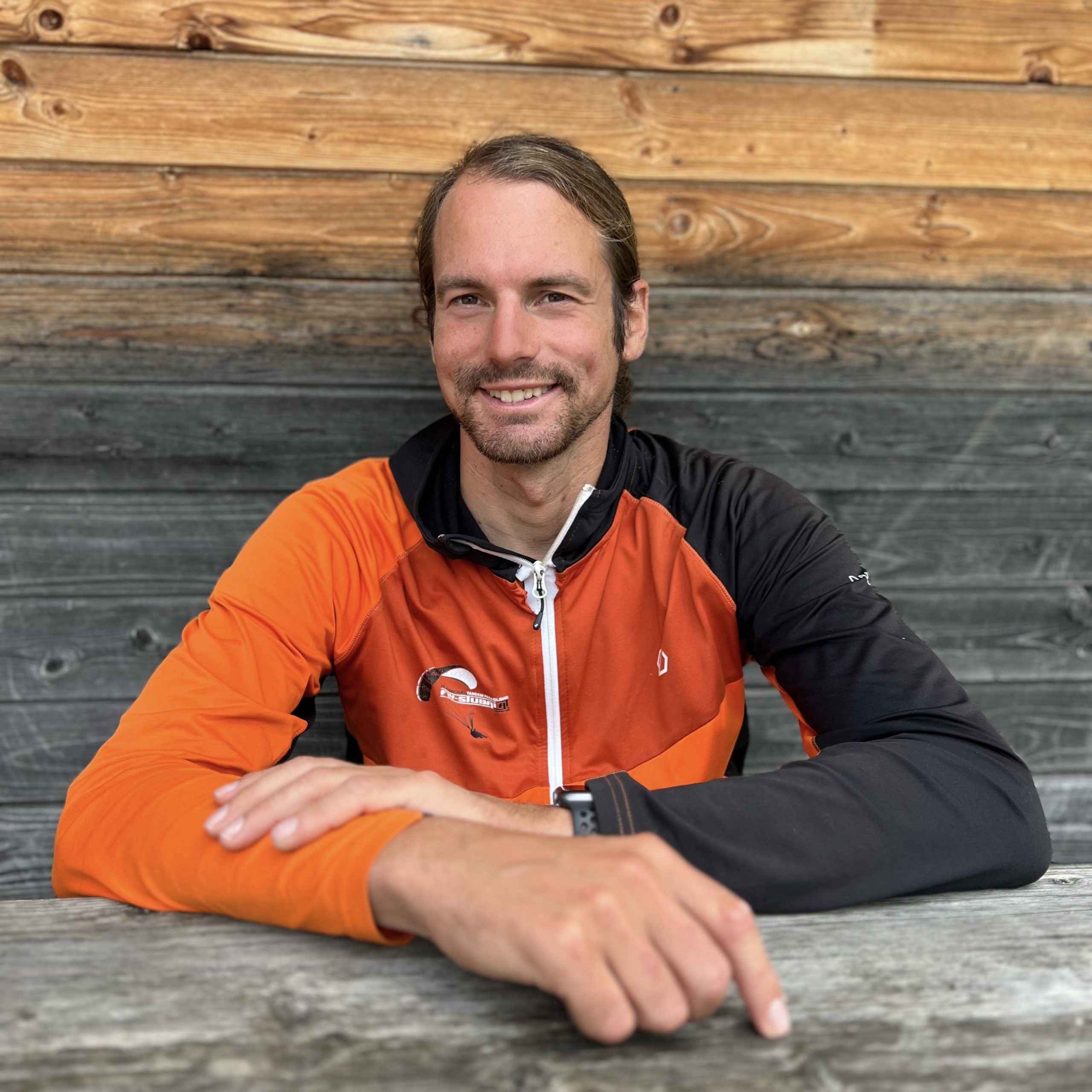 Ein Mann mit langen Haaren, der eine orange-schwarze Jacke trägt, sitzt an einem Holztisch und lächelt in die Kamera. Der Hintergrund besteht aus Holzbrettern in verschiedenen Farbtönen.