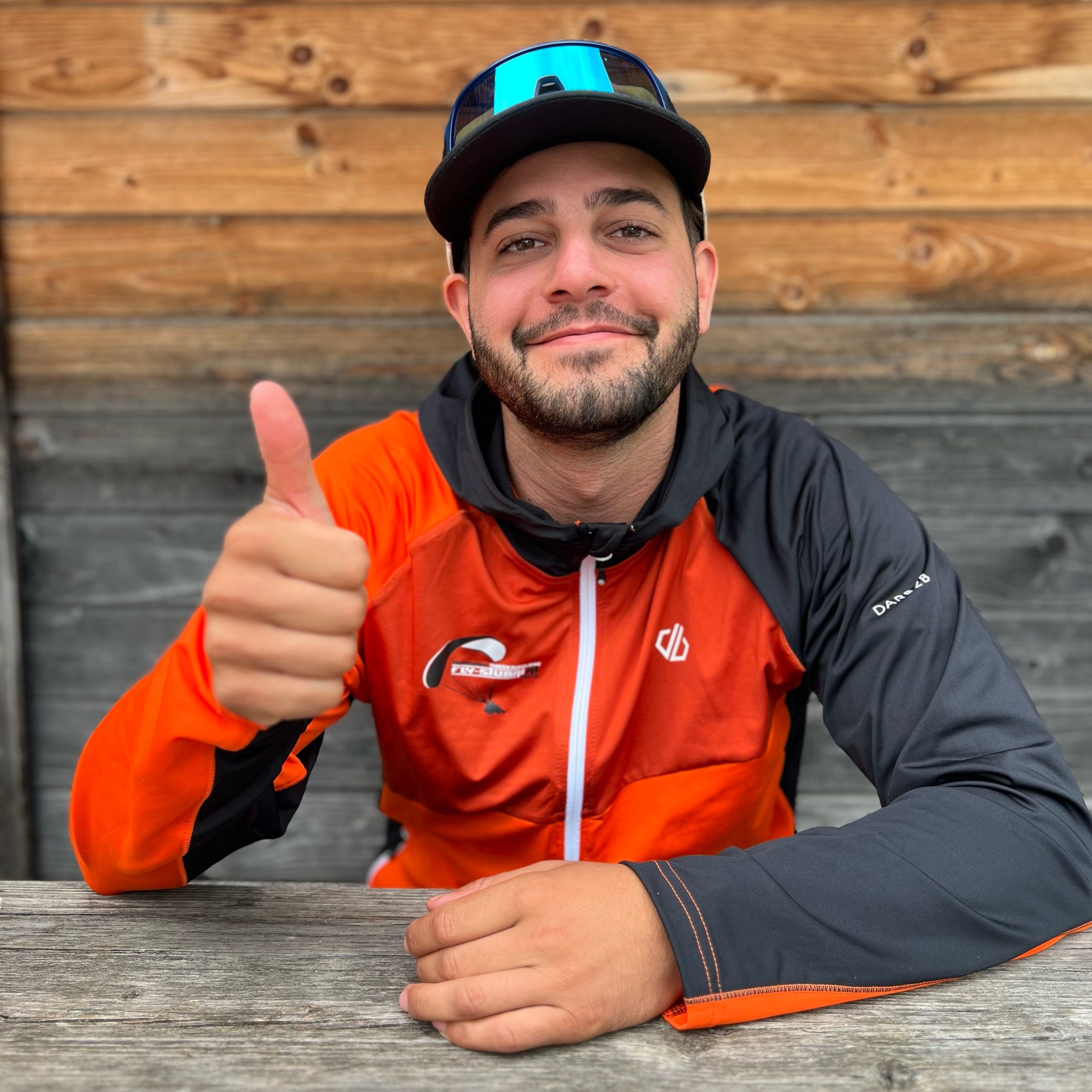 Ein Mann in einer orange-schwarzen Jacke sitzt an einem Holztisch, lächelt und zeigt den Daumen hoch. Er trägt eine Mütze und eine Sonnenbrille, im Hintergrund ist eine Holzwand zu sehen.